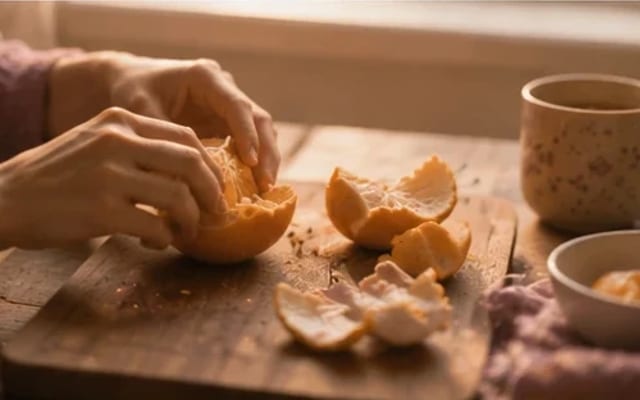 Mãos de mãe descascando uma laranja à luz quente — graça no lugar da culpa, amor no que não se vê